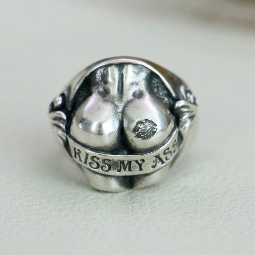Front-facing product shot of the silver sculpted butt signet ring on a light neutral background, displaying the "KISS MY ASS" engraved banner and lip mark accent