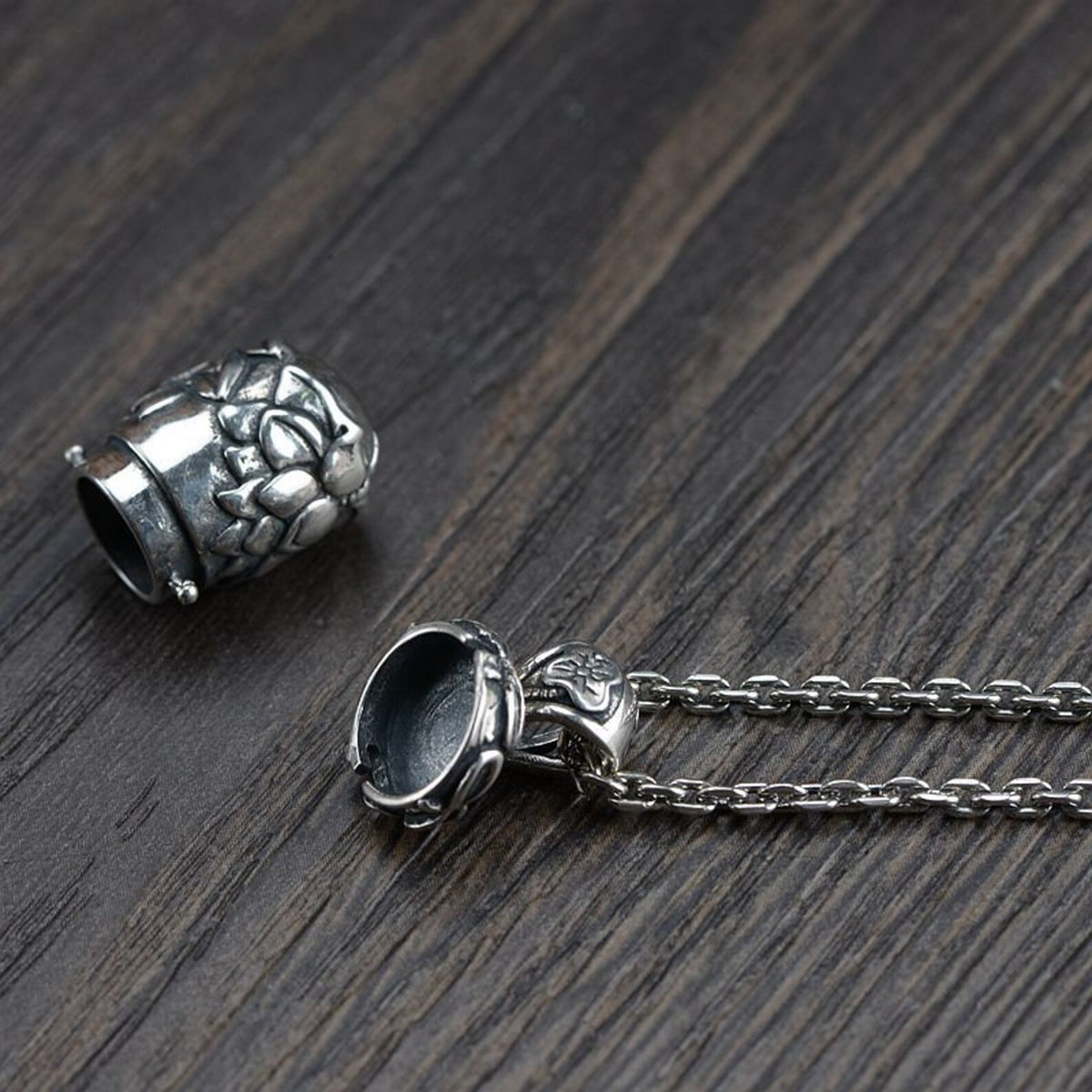 The sterling silver lotus locket pendant shown separated into its two main parts (the main body and the screw-top cap with the bail), lying on a dark wood surface