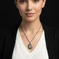 A woman wearing a silver pendant necklace with a circular medallion pendant, styled with a white V-neck top and a black cardigan against a dark background.