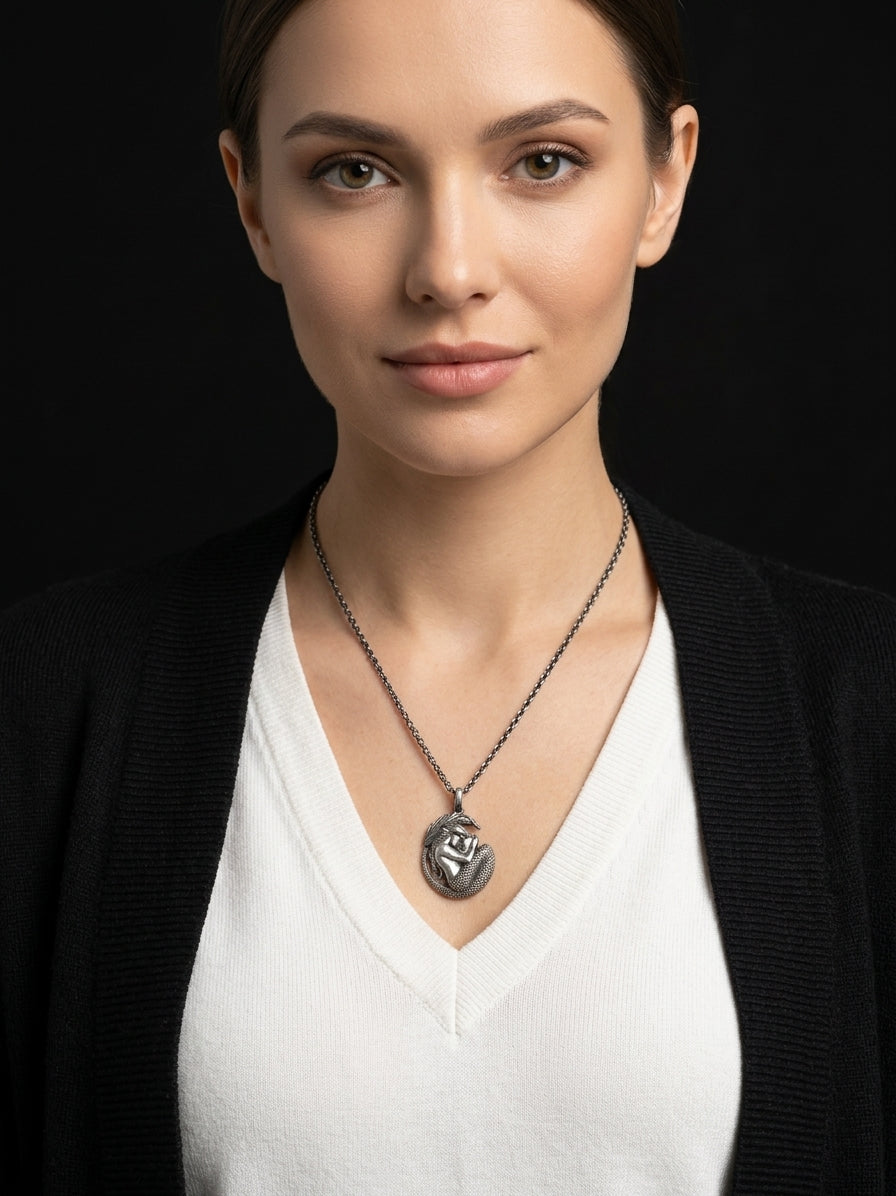 A woman wearing a silver pendant necklace with a circular medallion pendant, styled with a white V-neck top and a black cardigan against a dark background.