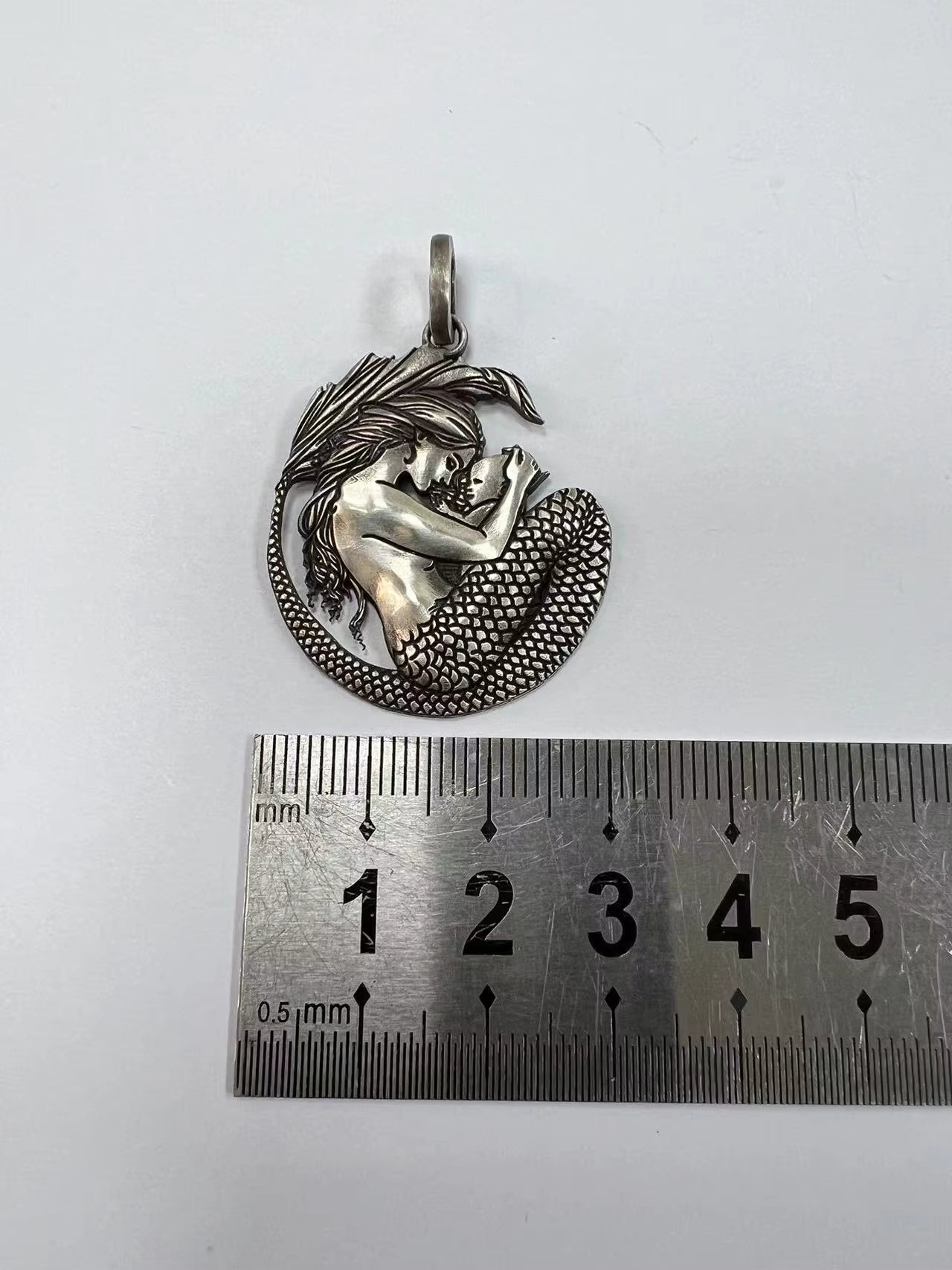 A silver pendant featuring a mermaid curled inside a shell, displayed above a metal ruler to indicate its size on a plain white background.